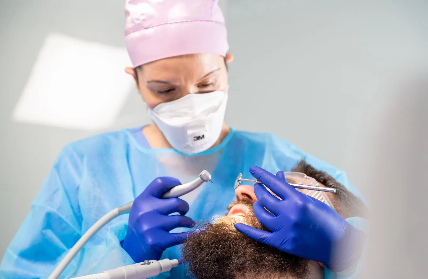 Dr. Roxana Usturoi, cu mască și halat de protecție, tratează un pacient folosind piesa de mână și oglinda dentară
